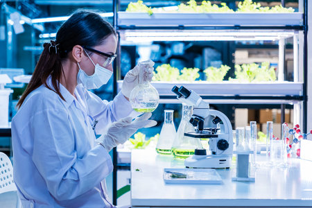 Female scientist examines green solution in a lab surrounded by plants and test tubes, representing eco-friendly farming innovations, sustainable agriculture, and plant-based biotechnology research.の写真素材