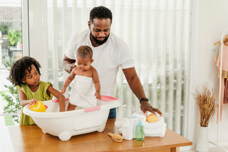 Father bathing baby in a small tub with help from older daughter, capturing authentic family care and diversity. A warm, candid moment of love, hygiene, and bonding in a modern home setting.の写真素材