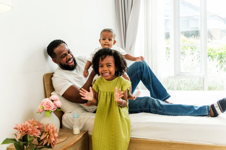 Joyful Black father plays with his baby and daughter on a bright bed in a cozy home, celebrating diverse family love and Fatherâs Day. A candid moment full of laughter, bonding, and parenthood joy.の写真素材