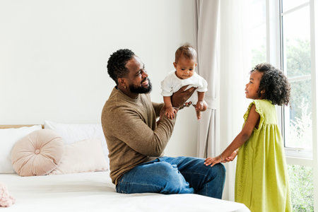 Joyful Black father holding baby while talking with his daughter near a window in a cozy bedroom. A warm, diverse family moment celebrating fatherhood, love, and authentic everyday parenting.の写真素材