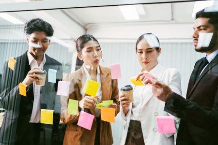 Multicultural office team collaborates in a brainstorming session, placing sticky notes on a glass wall. Business planning, creativity, and teamwork in a modern work environment with coffee in hand.の写真素材