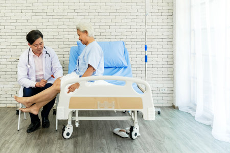 A male doctor conducts a knee reflex test on a senior woman sitting on a hospital bed. The scene highlights professional medical care, elderly wellness, and trust in a bright, clinical setting.の写真素材