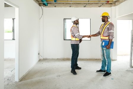 Two construction workers wearing safety gear and helmets shake hands inside a building under construction, symbolizing teamwork, real estate development, and success in the housing industry.の写真素材