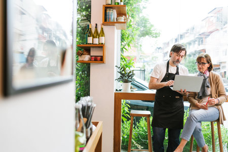 Cafe owner and financial advisor discussing business strategy using a laptop in a modern coffee shop. Bright setting with plants and wine bottles, symbolizing collaboration and entrepreneurship.の写真素材