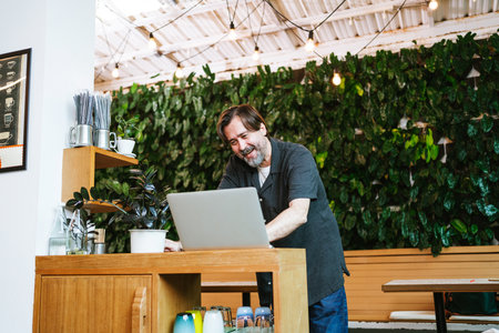Cheerful cafe owner chats with a financial advisor via video call at his cozy coffee shop. Surrounded by greenery, he discusses business strategies and growth opportunities with enthusiasm.の写真素材