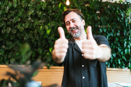 Happy middle-aged cafe owner giving two thumbs up in front of a lush green wall. He smiles confidently, celebrating business success and customer satisfaction in a cozy, eco-friendly setting.の写真素材