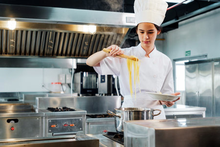 Young culinary student in a white chef uniform plating hot pasta from a steaming pot in a professional kitchen, demonstrating practical cooking skills during chef training at a culinary institute.の写真素材