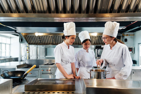 Chef instructing two culinary students in white uniforms and tall hats on how to cook in a commercial kitchen, standing near a pot on the stove during a hands-on professional cooking class.の写真素材