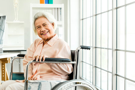 Senior woman smiling confidently in a wheelchair by a bright window in a clinic, representing independence, positive aging, rehabilitation, and elderly healthcare services with dignity and care.の写真素材