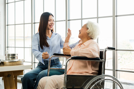 Young caregiver interacts warmly with a happy senior woman in a wheelchair near large windows, representing supportive elderly care, companionship, and assisted living in a healthcare environment.の写真素材