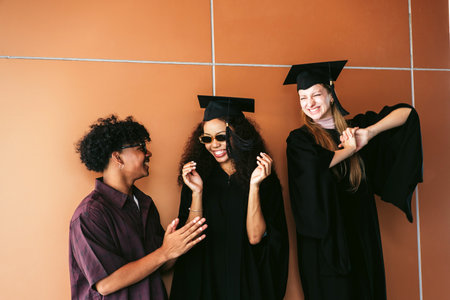 Joyful graduates in caps and gowns celebrate with laughter and applause. Diverse young friends share a cheerful moment after the ceremony, symbolizing success, pride, and friendship.の写真素材