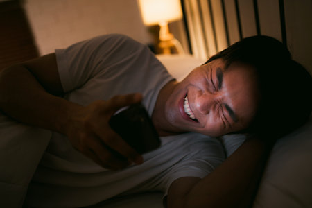 Young man lying in bed at night, laughing while looking at his smartphone screen. Concept of digital entertainment, bedtime phone use, relaxation, and screen time before sleep.の写真素材