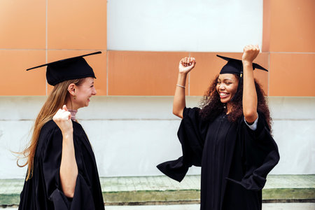 Two joyful young female graduates raise their arms in celebration, sharing a moment of triumph and success outdoors in graduation gowns and caps. A proud and cheerful milestone achievement moment.の写真素材