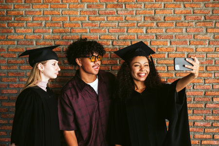 Happy diverse graduates celebrate with a selfie against a brick wall. Two students in caps and gowns smile with a friend, capturing joy and pride on graduation day. Friendship and achievement concept.の写真素材