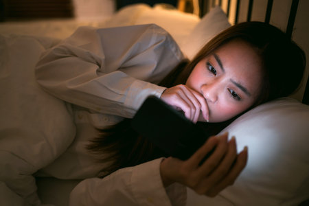 Young woman lying in bed late at night, staring at smartphone screen with concerned expression. Concept of sleep disruption, eye strain, and mental health impact from late-night mobile device use.の写真素材