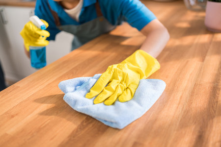 Close-up of a professional cleaner wiping a wooden kitchen counter with a blue microfiber cloth and spray bottle, emphasizing hygiene, sanitation, and premium home cleaning service standards.の写真素材