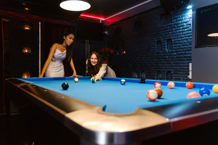 Two stylish women enjoy a friendly game of pool at a modern lounge. One lines up a shot while the other cheers on, capturing nightlife fun, friendship, and leisure in a trendy bar setting.の写真素材
