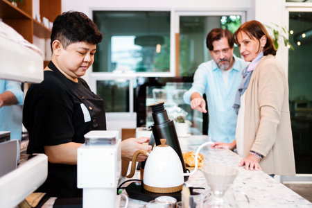 Cafe barista prepares coffee while couple discusses drink order across counter. Small business service scene highlighting friendly customer interaction in a cozy, modern shop environment.の写真素材