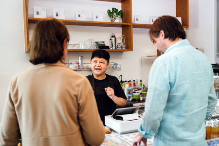 Cafe owner explains menu to customers at counter in modern coffee shop. Small business engagement, friendly service, and hospitality in a welcoming local setting with diverse interaction.の写真素材