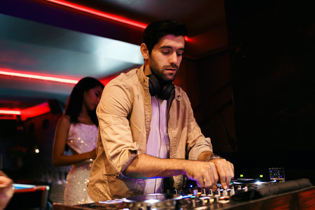 Male DJ focused on mixing music at nightclub, wearing headphones and casual jacket. Vibrant red lighting sets party mood, with background club goers enjoying the lively evening scene.の写真素材