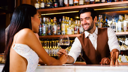 Smiling bartender chats with a stylish woman holding a wine glass at an elegant bar. Warm lighting and shelves of bottles set a friendly, upscale nightlife scene with relaxed, social vibes.の写真素材