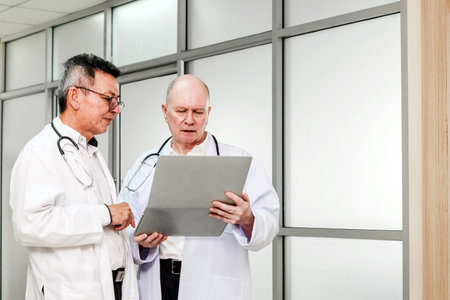 Two senior male doctors in lab coats discussing a patient's case while reviewing information on a digital tablet. Professional collaboration in a modern hospital setting, focused on medical teamwork.の写真素材
