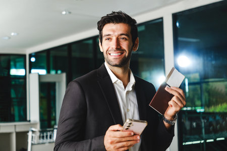 Smiling businessman holding smartphone, passport, and boarding pass at the airport. Concept of air travel, check-in service, and professional global mobility with modern digital convenience.の写真素材