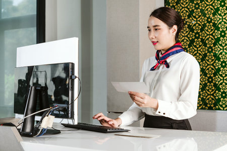 Airline ground staff woman in uniform assists check-in at airport counter, holding ticket and using computer. Concept of air travel service, passenger support, and airport operations.の写真素材
