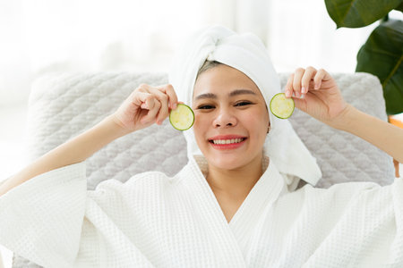 Happy woman in white bathrobe and towel turban holds cucumber slices near her eyes, smiling brightly. Represents skincare, beauty, spa wellness, and joyful self-care in a serene home spa setting.の写真素材