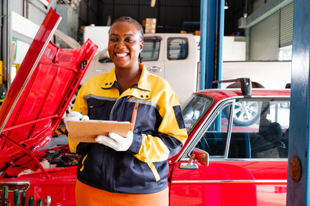 Smiling female auto technician holding a clipboard in a garage beside a red car with its hood open. Represents confidence, professionalism, and diversity in automotive repair services.の写真素材