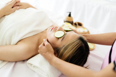 Woman enjoys a facial spa treatment with cucumber slices on her eyes while lying under a white towel. A serene wellness setting promoting skincare, relaxation, and natural beauty therapy.の写真素材