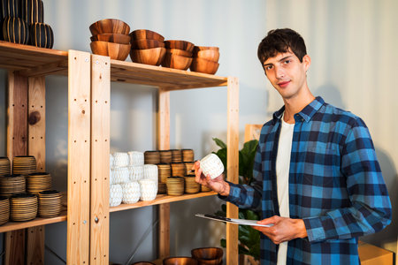 Young man in plaid shirt holding a ceramic cup and clipboard while standing near wooden shelf with...