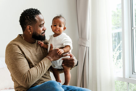 Loving father holds his baby girl near a bright window, sharing a tender moment. An authentic diversity family portrait symbolizing fatherhood, care, bonding, and Fatherâs Day celebration.の写真素材