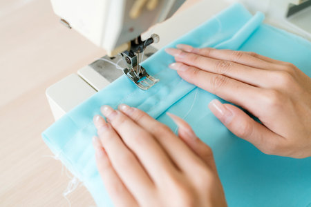 Closeup of hands stitching blue fabric under sewing machine in bright studio. Seamstress at work...