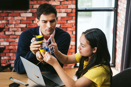 Two coworkers in modern workshop collaborate on robotics prototype at laptop. Engineering design,...