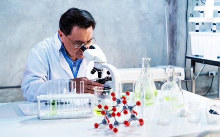 Scientist using a microscope at a lab bench with test tubes, flasks, and a molecular model, representing laboratory research, analysis, and innovation in modern science.の写真素材