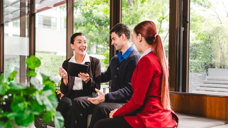 Three business professionals meet in modern hotel lobby, discussing ideas on tablet. Scene shows collaboration, consulting, client partnership, negotiation, teamwork, and presentation planning.の写真素材