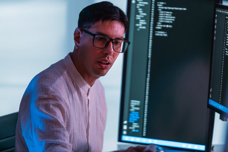 Programmer wearing glasses codes at dual monitors in modern office, lit by cool blue light. Scene highlights developer focus, software engineering, technology, and computer workstation setup.の写真素材
