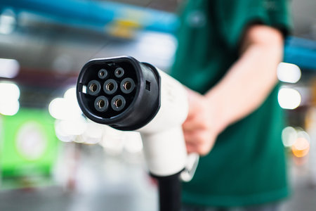 Close-up of a person holding an electric vehicle charging plug, with blurred lights in the...