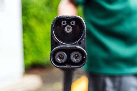 Close-up of an electric vehicle charging connector held outdoors, showing charging pins and cable,...