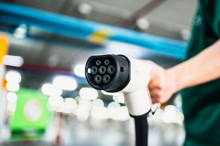 Person holding an electric vehicle charging plug in a blurred indoor setting, showing the connector...