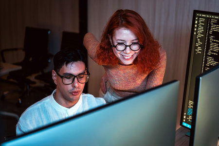 Two coworkers collaborate at dual monitors in a dim office, reviewing code and brainstorming...