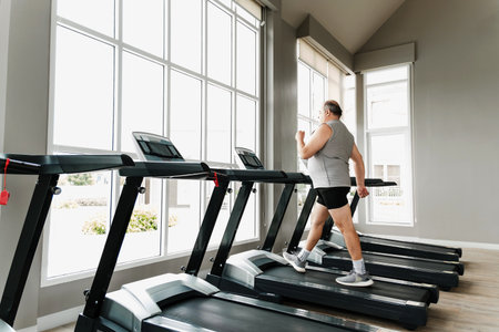 Man walks on treadmill in bright, spacious gym with large windows. Scene shows cardio fitness,...
