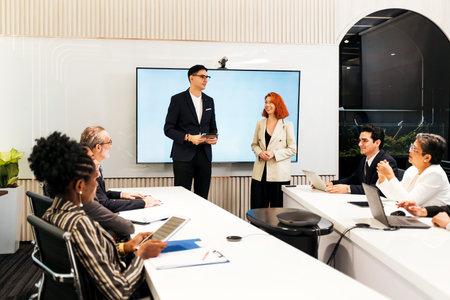 Diverse team in a modern conference room during a presentation. Two colleagues stand by a large...