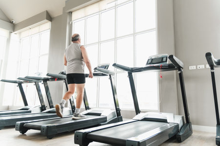 Man walking on a treadmill in a bright modern gym with large windows, multiple machines, and natural...