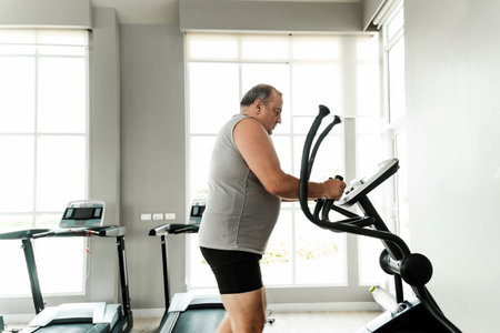 Senior man trains on elliptical in bright gym with large windows, sweating through cardio workout. Scene shows strength, motivation, discipline, rehabilitation, recovery, effort, and overall wellness.の写真素材