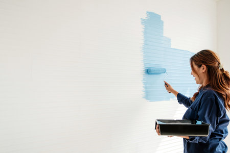 Woman painting a white interior wall light blue with a roller, holding a paint tray during a home renovation project, with ample copy space on the textured surface.の写真素材