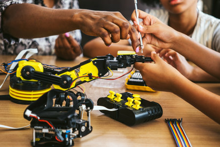 Hands collaborating to assemble a yellow robotic arm kit on a workbench, using tools and wires during a STEM workshop, illustrating teamwork, engineering, and practical robotics education.の写真素材