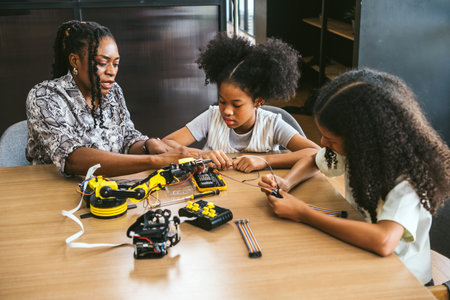 Teacher guides two girls building a robotics kit at a classroom table, assembling circuits and...