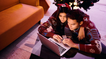 Couple in festive sweaters shopping online at home by a Christmas tree, using a laptop and credit card on a glass table, cozy holiday evening atmosphere.の写真素材